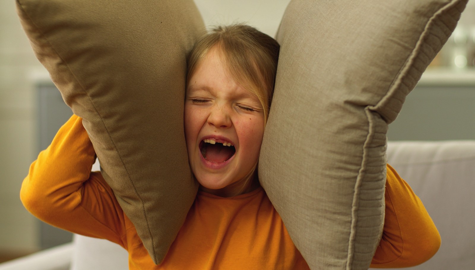 Ein Kind in orangefarbenem Pullover presst sich schreiend zwei Sofakissen an die Ohren.